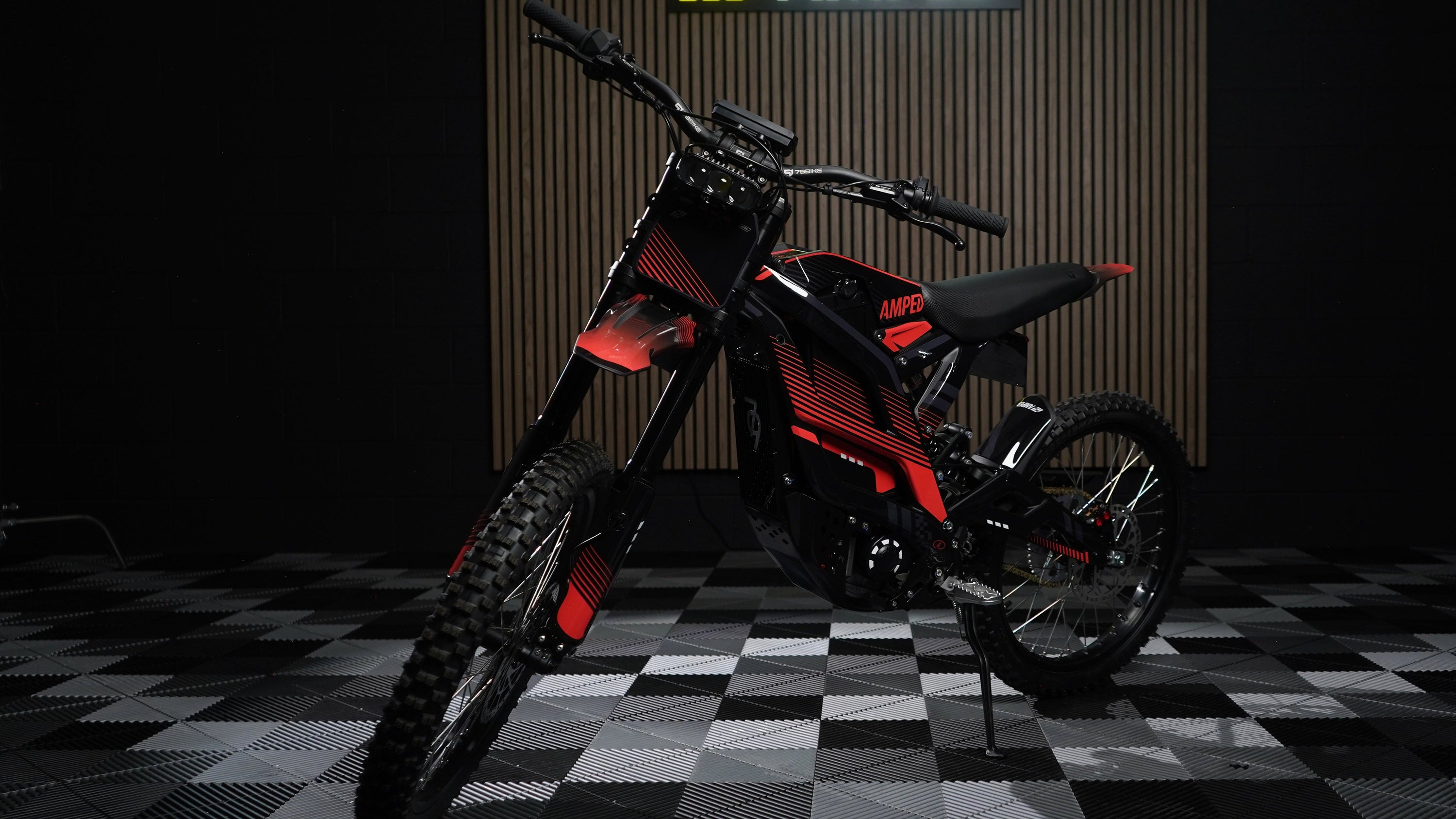 A red and black graphic Falcon GT electric dirt bike displayed on a stand in a studio setting with a checkered floor and a vertical wood-grain backdrop featuring the AMPED logo.