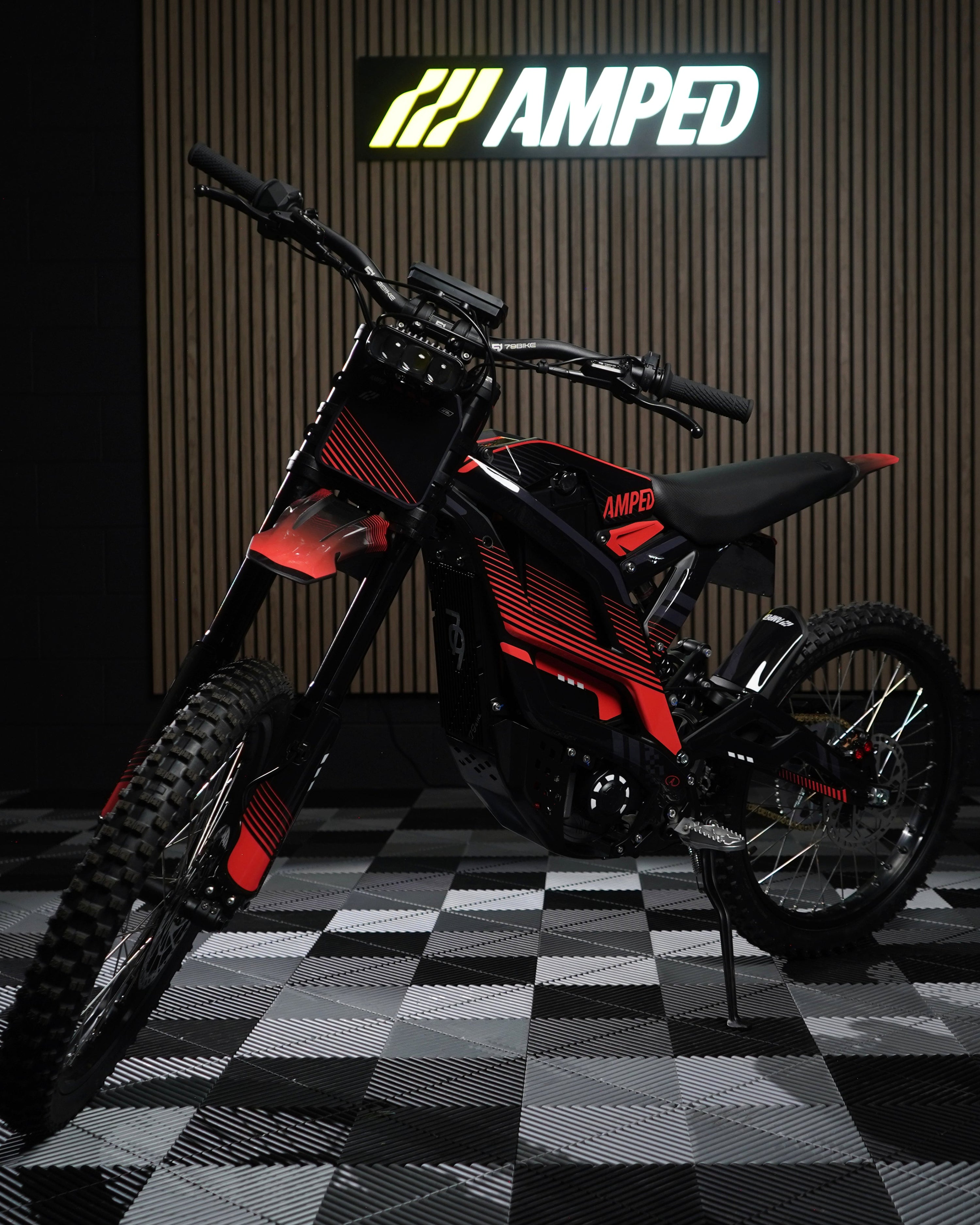 A red and black graphic wrap on a 79BIKE Falcon electric dirt bike displayed on a stand in a studio setting with a checkered floor and a vertical wood-grain backdrop featuring the AMPED logo.
