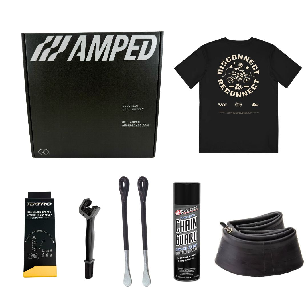 On a white background, there is an AMPED box containing a black T-shirt with DISCONNECT RECONNECT graphic and the Essential E-Moto Care Kit by Amped Bikes, featuring Tektro brake pads, bike tire levers, Amped chain guard spray, and a bicycle inner tube.