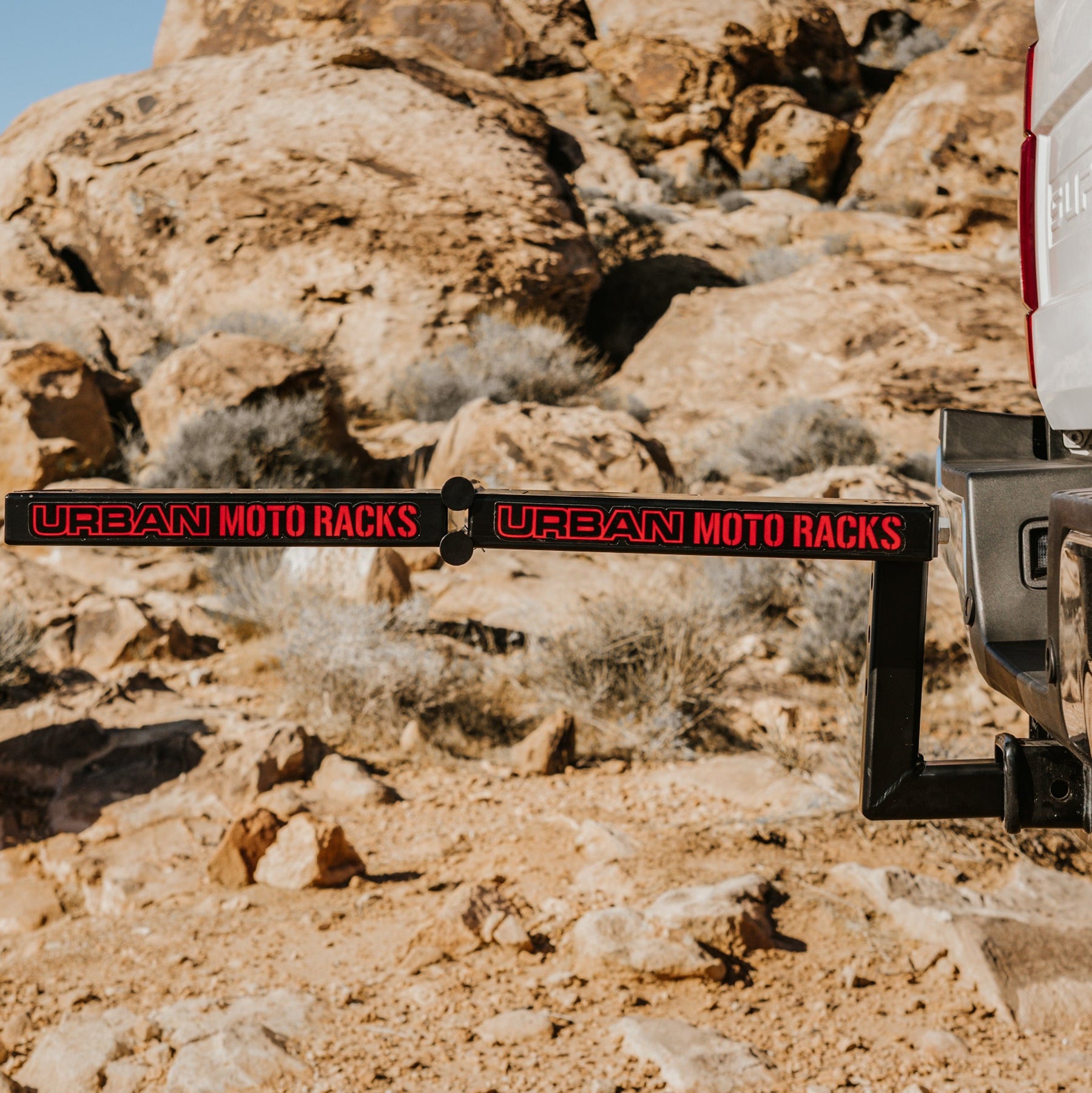 A sleek Urban Moto Racks by Amped Bikes is mounted on a vehicle, designed for E-Moto models, against a sunlit desert landscape with large rocks in the background.