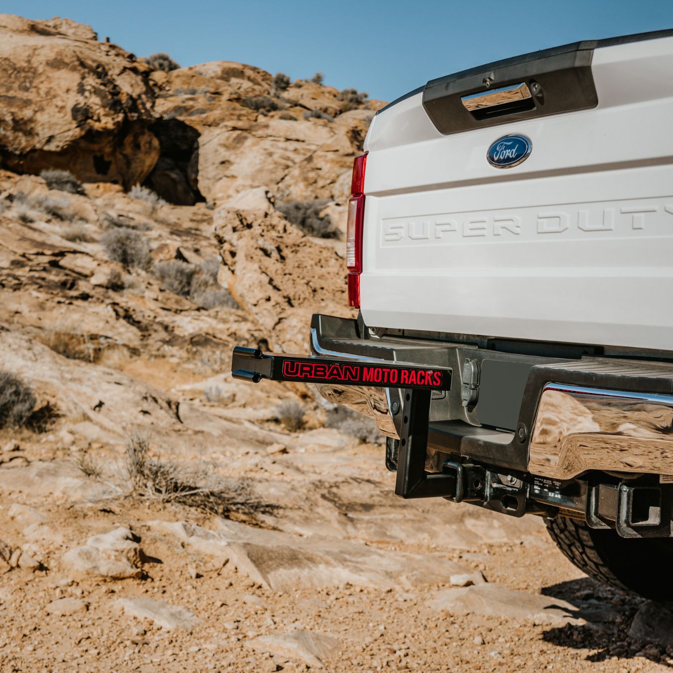 A white Ford Super Duty truck, parked in a rocky desert with sparse vegetation, features a red and black Urban Moto Rack by Amped Bikes, specifically designed for E-Moto models.