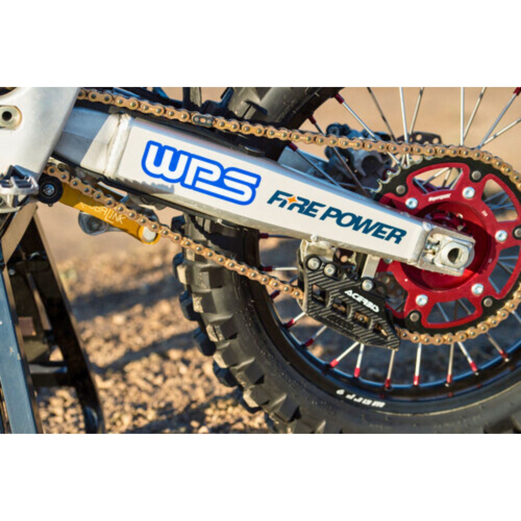 Close-up of a dirt bikes rear wheel featuring the Fire Power Heavy Duty Chain 428X120 Gold, with detailed shots of the chain, sprocket, and swingarm. The Amped Bikes brand stands out against a blurred dirt terrain background—perfect for fans of the sturdy Talaria Sting MX5 Pro.