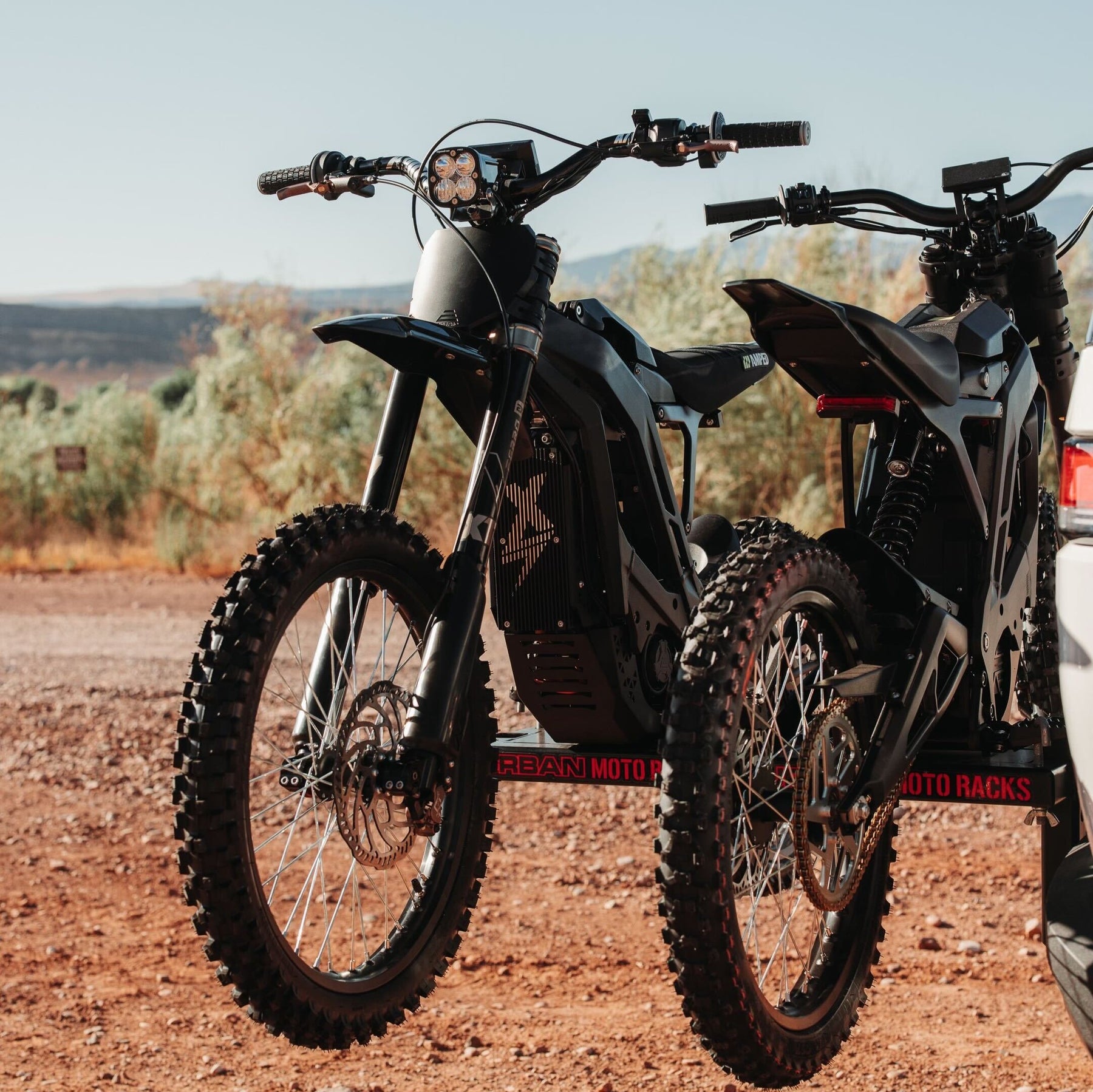 Two off-road motorcycles with knobby tires are mounted on Amped Bikes Urban Moto Racks at the back of a vehicle in a desert landscape. The sun casts shadows, highlighting the rugged terrain and sparse vegetation.