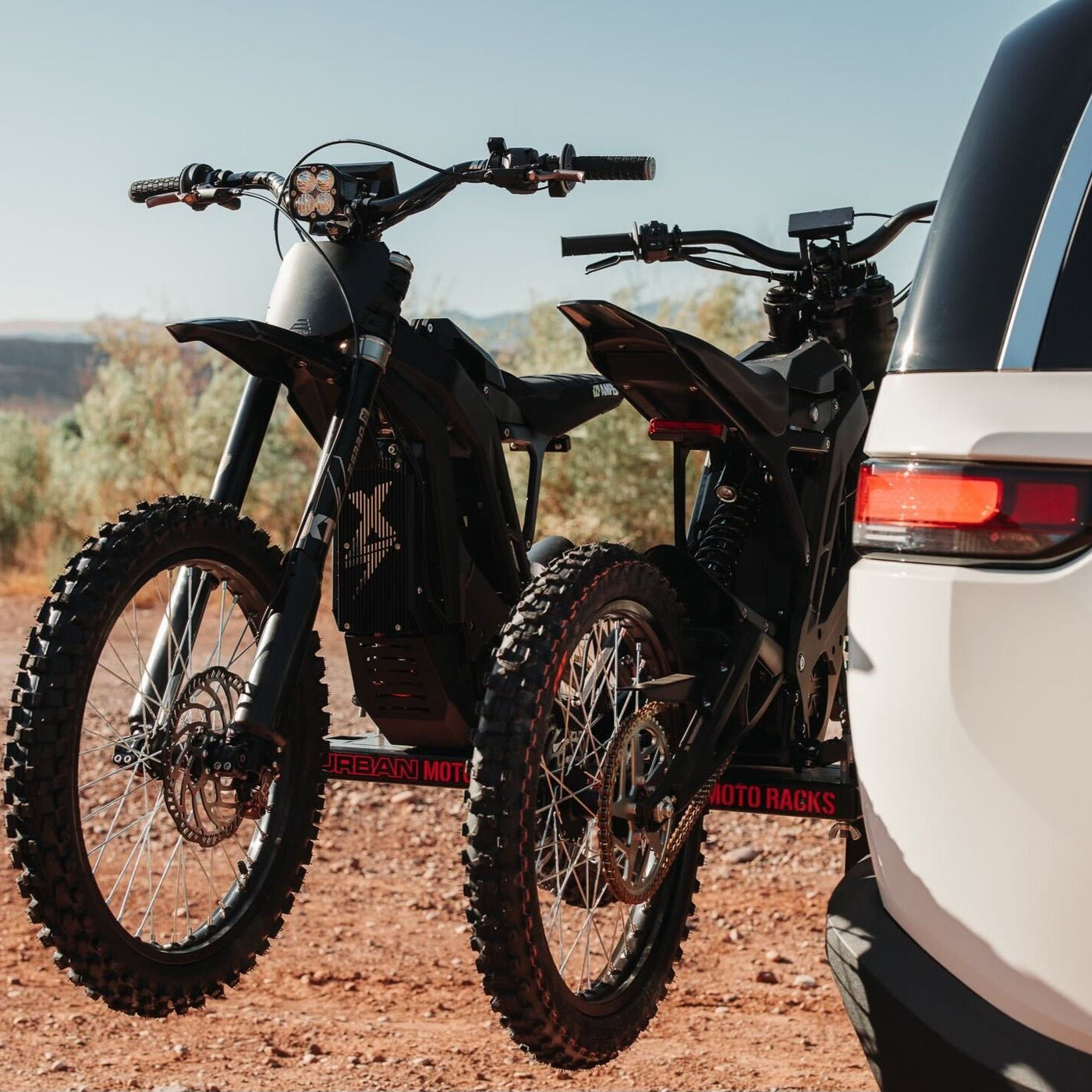 Under a clear sky, surrounded by trees and reddish dirt, two electric bikes are mounted on Amped Bikes Urban Moto Racks at the rear of a white vehicle, perfectly blending nature and technology.