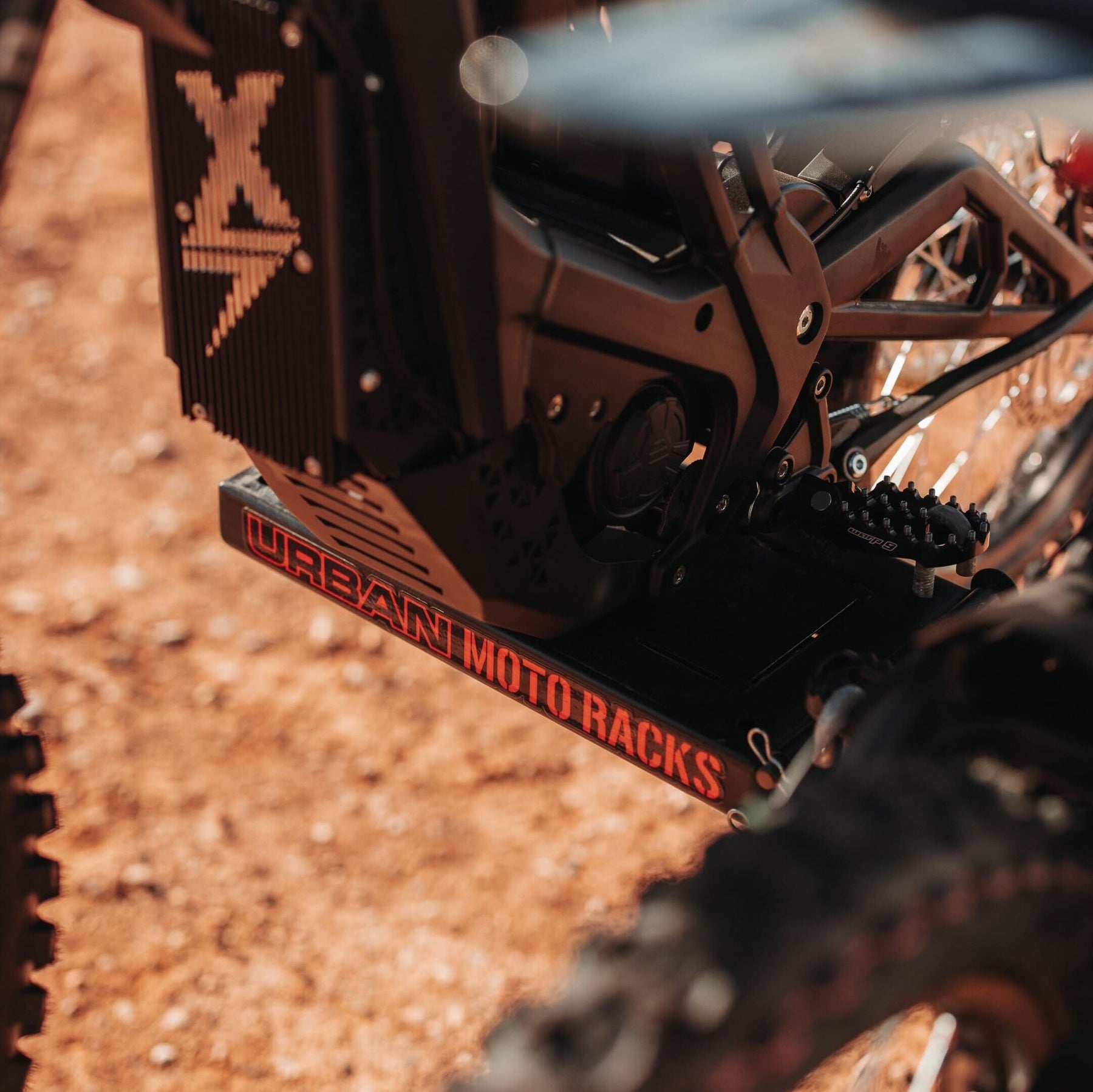 Close-up of a motorcycle featuring a rugged, black frame with the Urban Moto Racks name highlighted in bold red lettering. Set against a textured, earthy background, the scene exemplifies Amped Bikes E-Moto models ready to tackle any terrain.