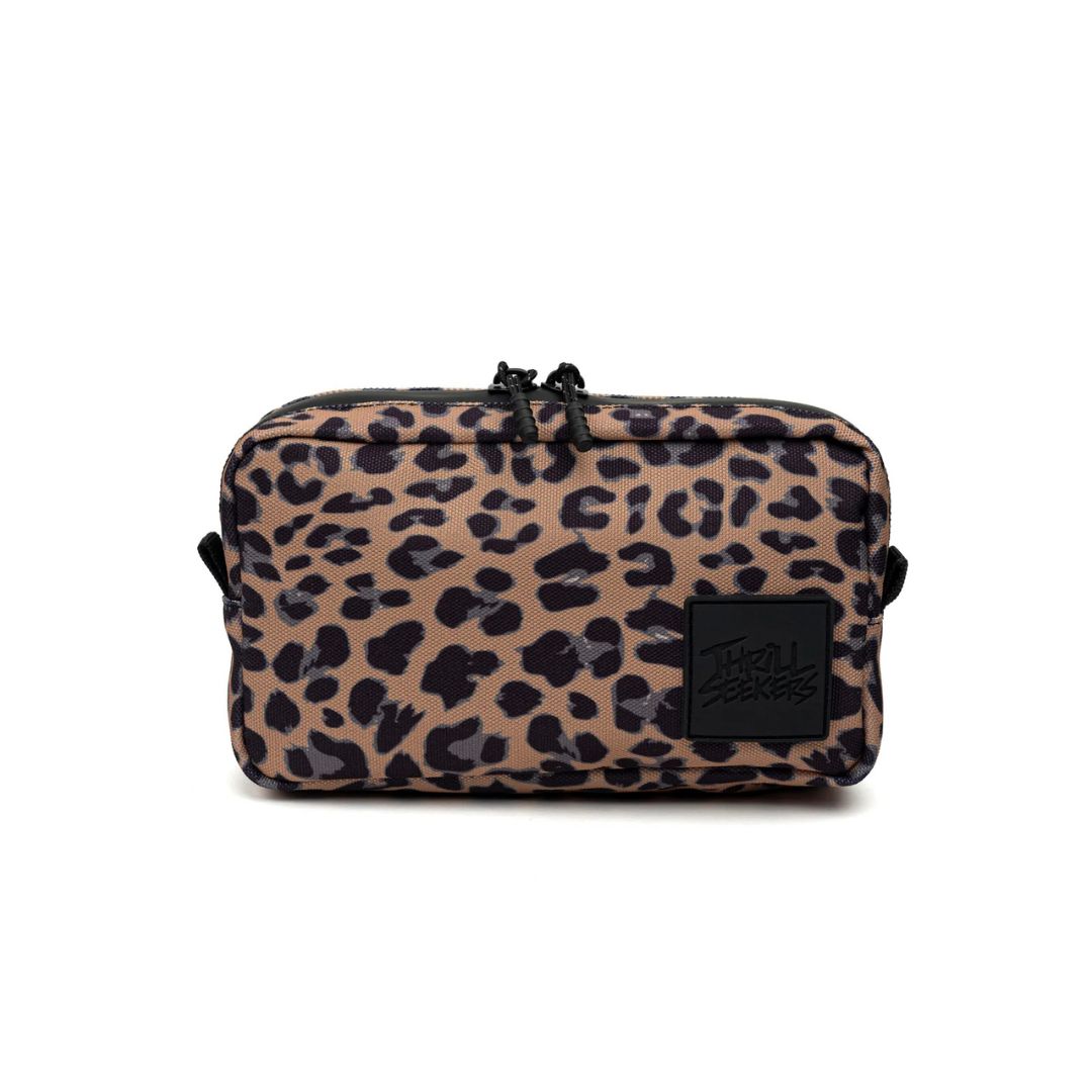 The Bar Bag V2 by Thrill Seekers is a rectangular bag with a leopard print and a front patch reading Thrill Seekers. It includes a water-resistant zipper to securely store essentials in its compact design.